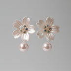 A pair of flower-shaped stud earrings with translucent pale pink petals, iridescent rhinestone centers, and dangling pearl-like drops, displayed on a pink card.