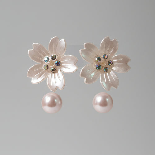 A pair of flower-shaped stud earrings with translucent pale pink petals, iridescent rhinestone centers, and dangling pearl-like drops, displayed on a pink card.